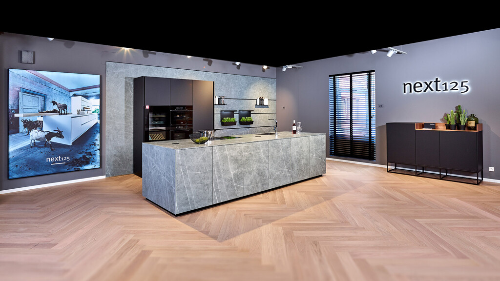 Blick auf eine Küche mit Insel in Betonoptik und dunklen Hochschränken in unserer Ausstellung von next125