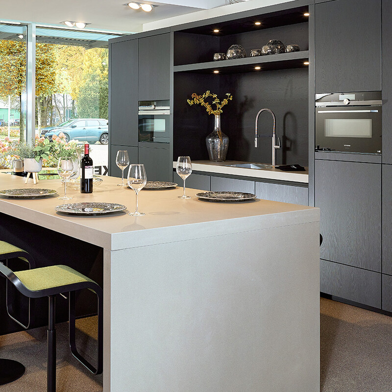 Diese Küche mit Nische in der Schrankwand ist eine schwarze Inselküche von Contur mit einer Keramikarbeitsplatte.