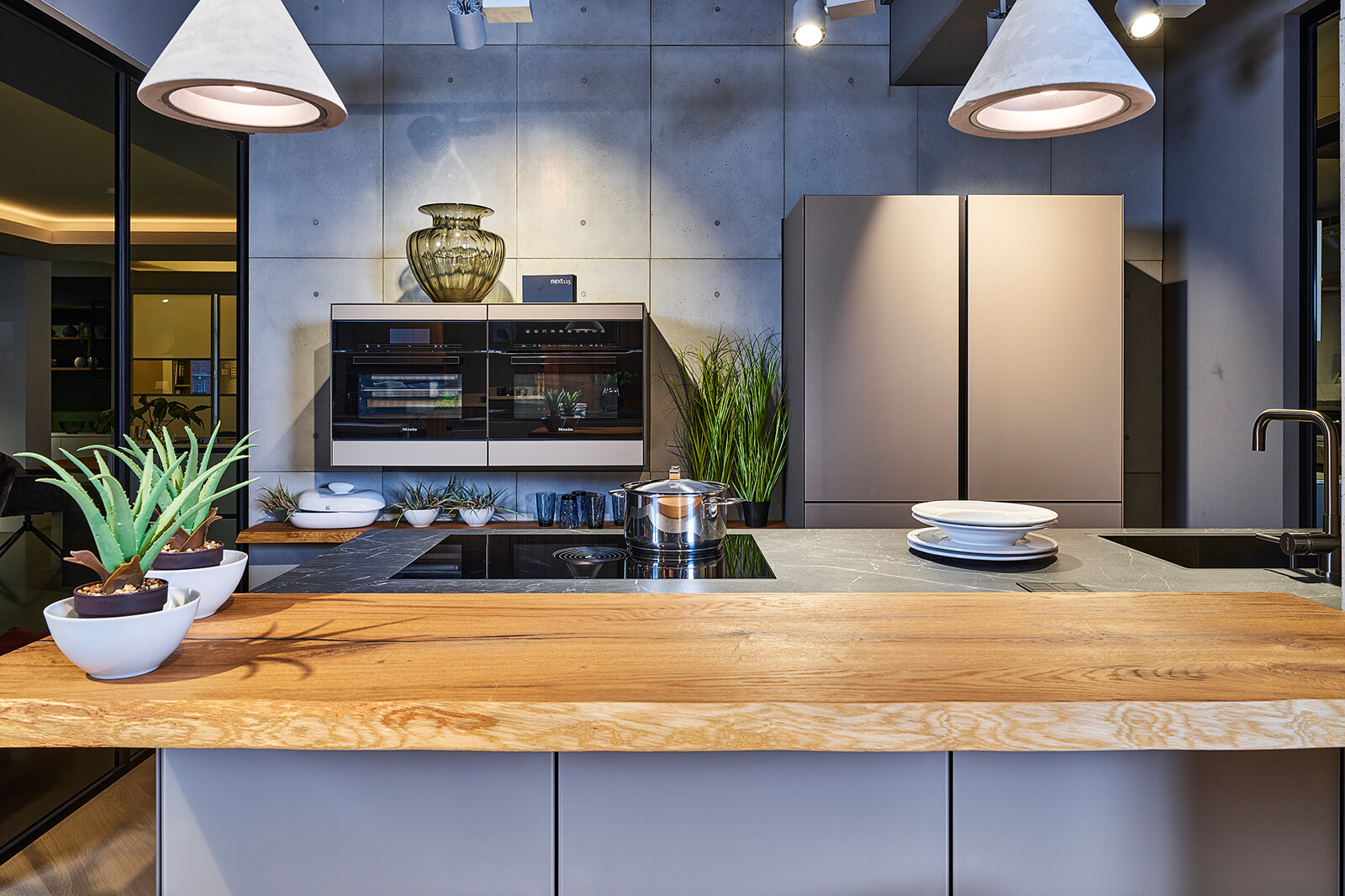 Showroomkeuken: next125 compacte industriële eilandkeuken met matglas fronten, houten bar en gaggenau ovens