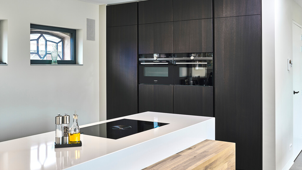 Bijzondere eilandkeuken die deels uit SieMatic S2 elementen bestaat en deels maatwerk uit onze eigen werkplaats is. De massief eiken ombouw met tafel om het witte SieMatic eiland, en de donkere eiken fronten van de hoge kasten zijn maatwerk.