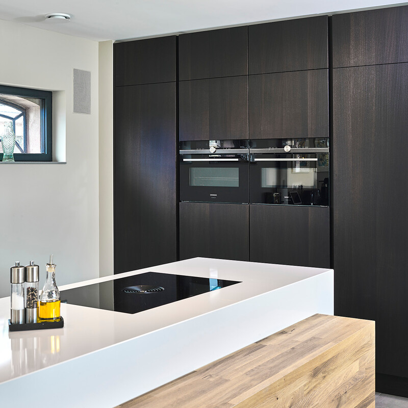 Bijzondere eilandkeuken die deels uit SieMatic S2 elementen bestaat en deels maatwerk uit onze eigen werkplaats is. De massief eiken ombouw met tafel om het witte SieMatic eiland, en de donkere eiken fronten van de hoge kasten zijn maatwerk.