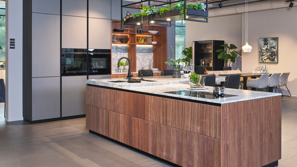 Außergewöhnliche Inselküche von next125 mit geriffelten Fronten in Nussbaum und einem Hochschrank mit Mattglasfronten in Basaltgrau. Mit Küchenarbeitsplatte aus Granit in der Ausführung Superwhite. Zu sehen beim Küchenland Ekelhoff in Nordhorn